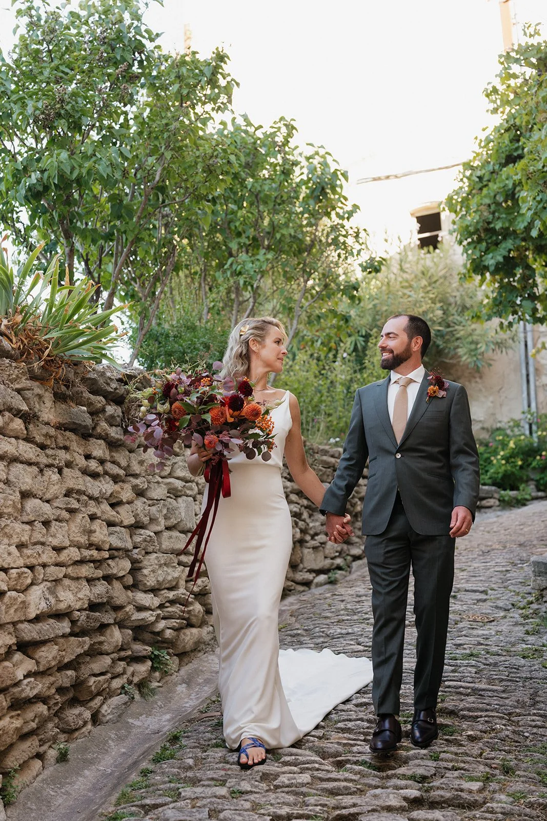 A bride and groom walking hand in hand along a cobblestone path, surrounded by green trees and plants. The bride holds a colorful bouquet of flowers, and both are smiling at each other.