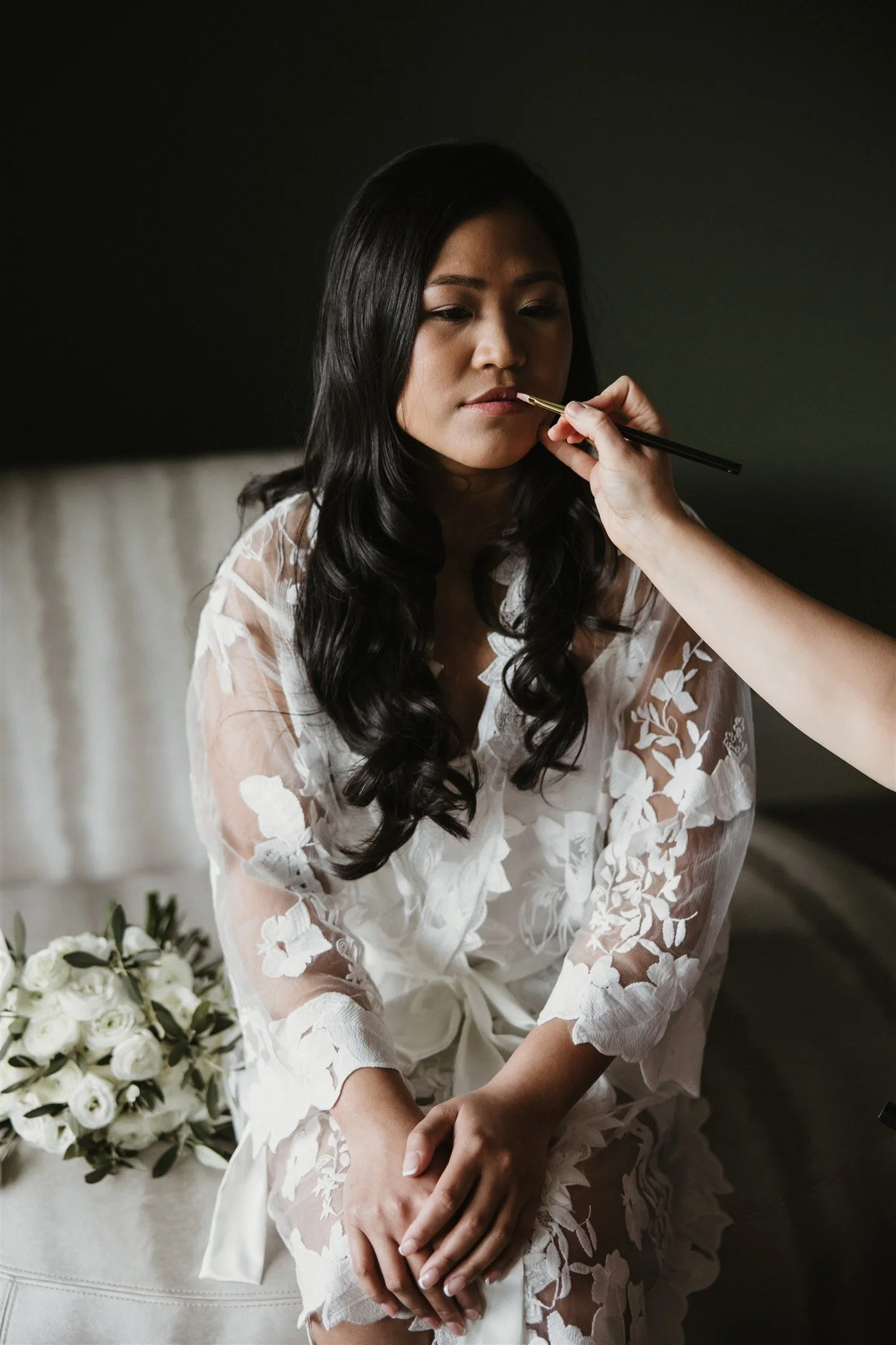 A young woman with long black hair in a white lace robe, sitting with her hands on her lap, having makeup applied by a person holding a brush. There is a bouquet of white roses on a table nearby.