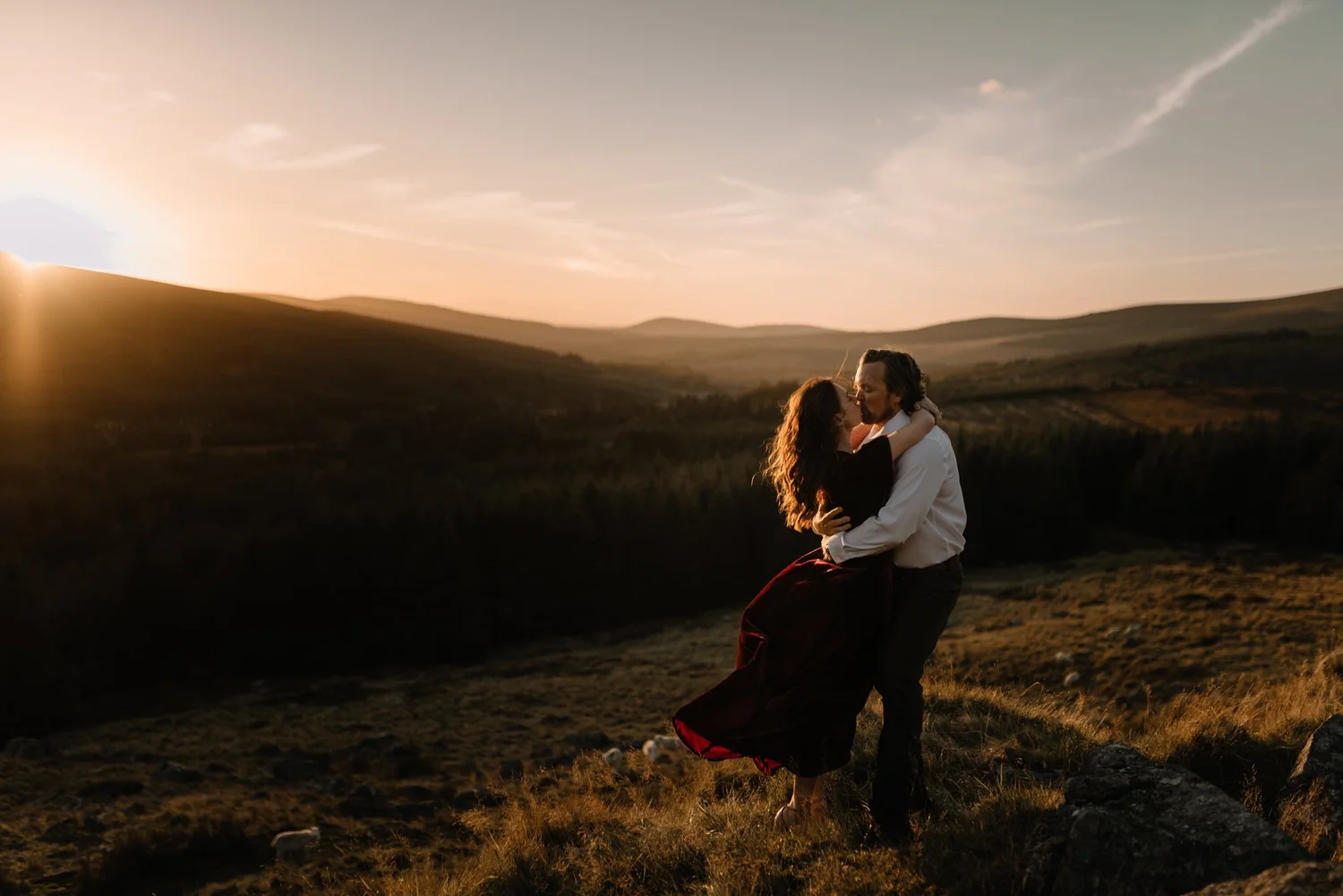 A couple embracing and kissing outdoors during sunset with rolling hills in the background.