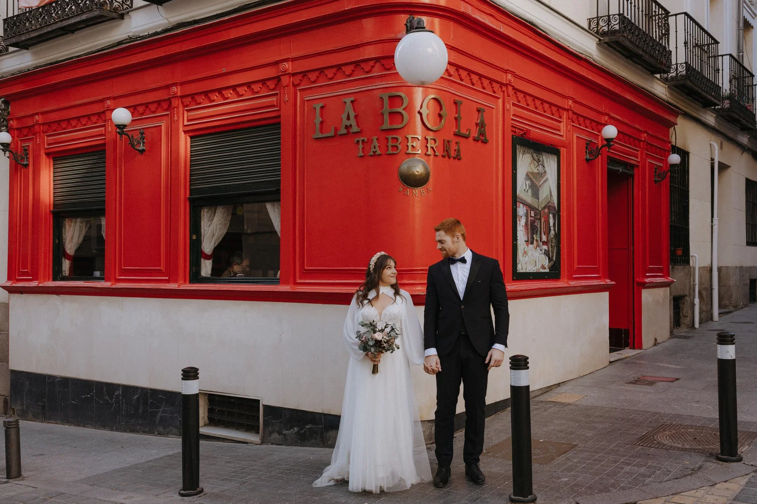 A bride and groom holding hands outside a red building named La Bolla Taberna, with the bride holding a bouquet of flowers and wearing a white dress and headband, and the groom wearing a black tuxedo and bow tie.