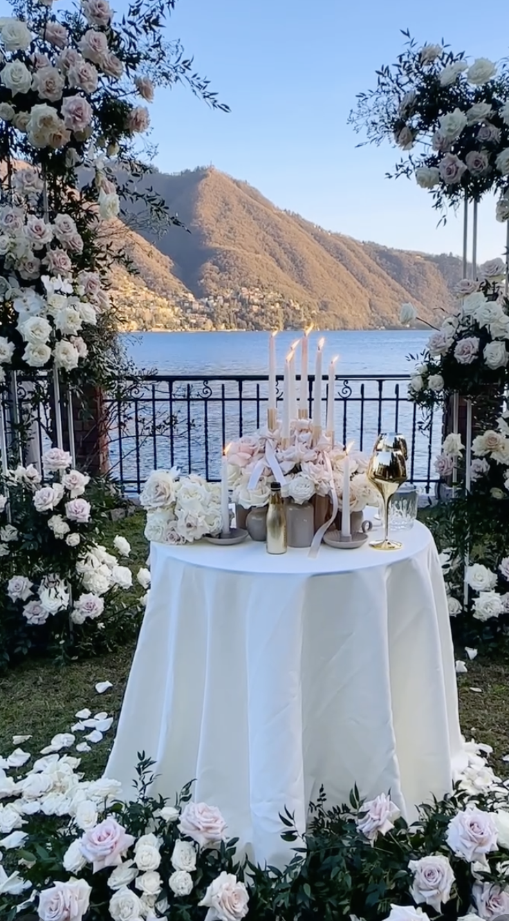 A wedding altar setup with white and pink roses, candles, and gold accents overlooking a lake with mountains in the background.