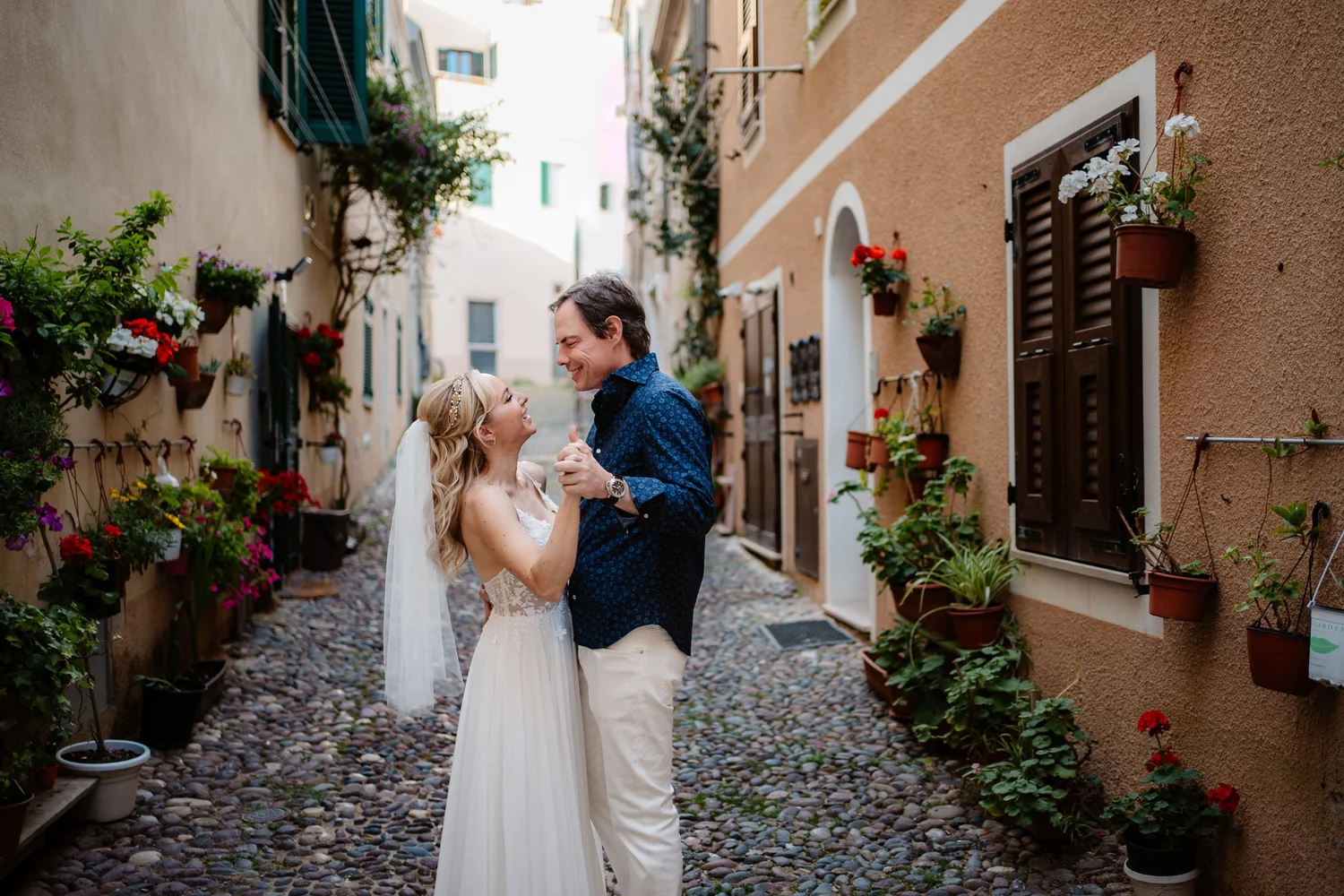 A bride and groom dancing in a narrow European-style alleyway lined with colorful potted flowers on the walls.