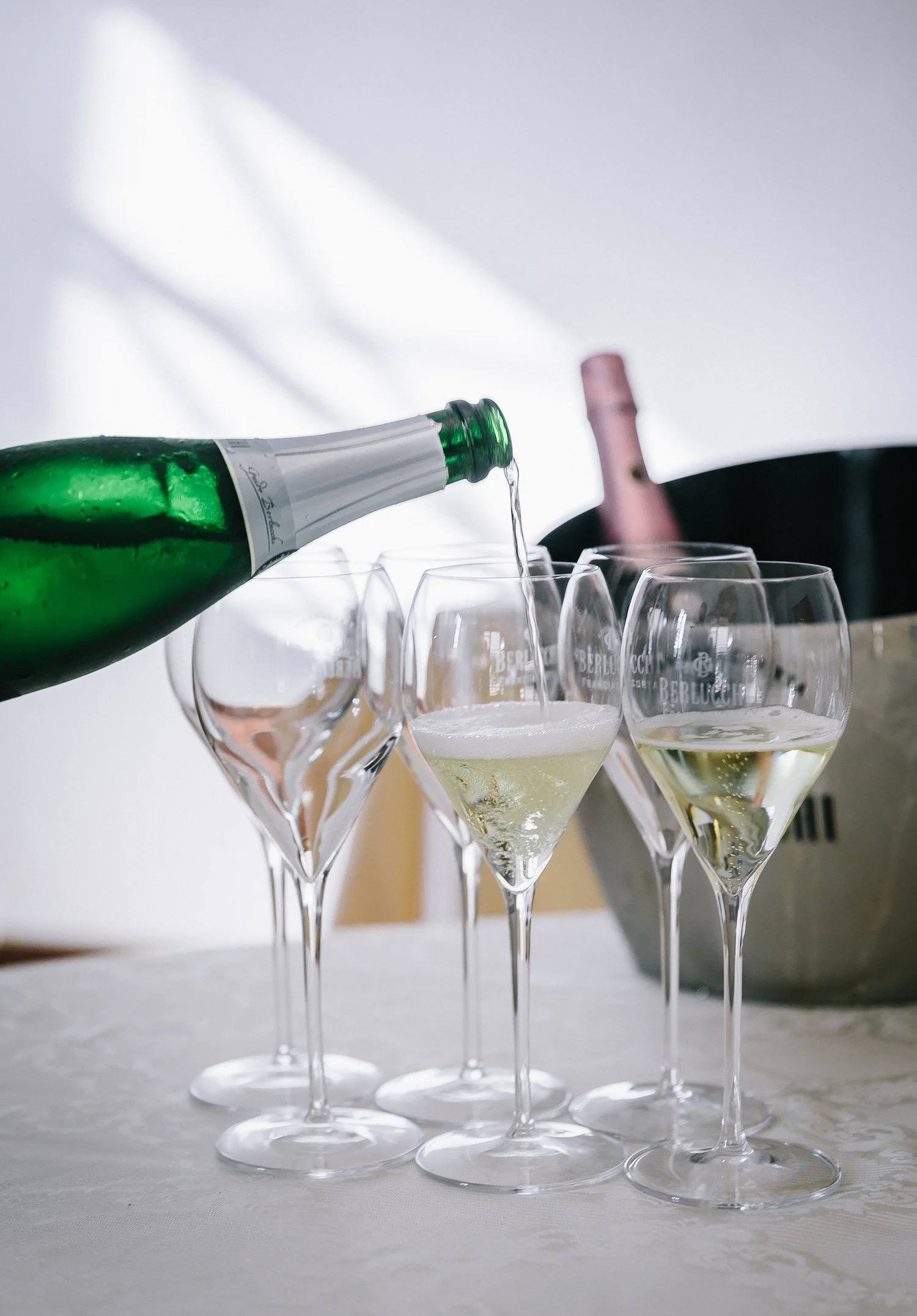 A green champagne bottle pouring into a glass, with several empty champagne glasses on a white tablecloth.