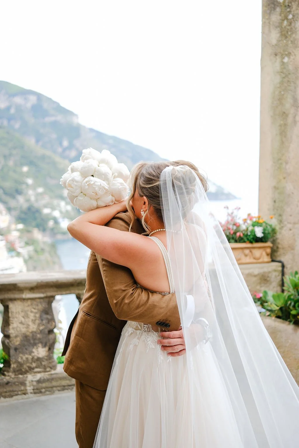 Bride and groom embrace during their wedding, with the bride wearing a white gown and veil and the groom in a brown suit, on a terrace with scenic mountain and water view.