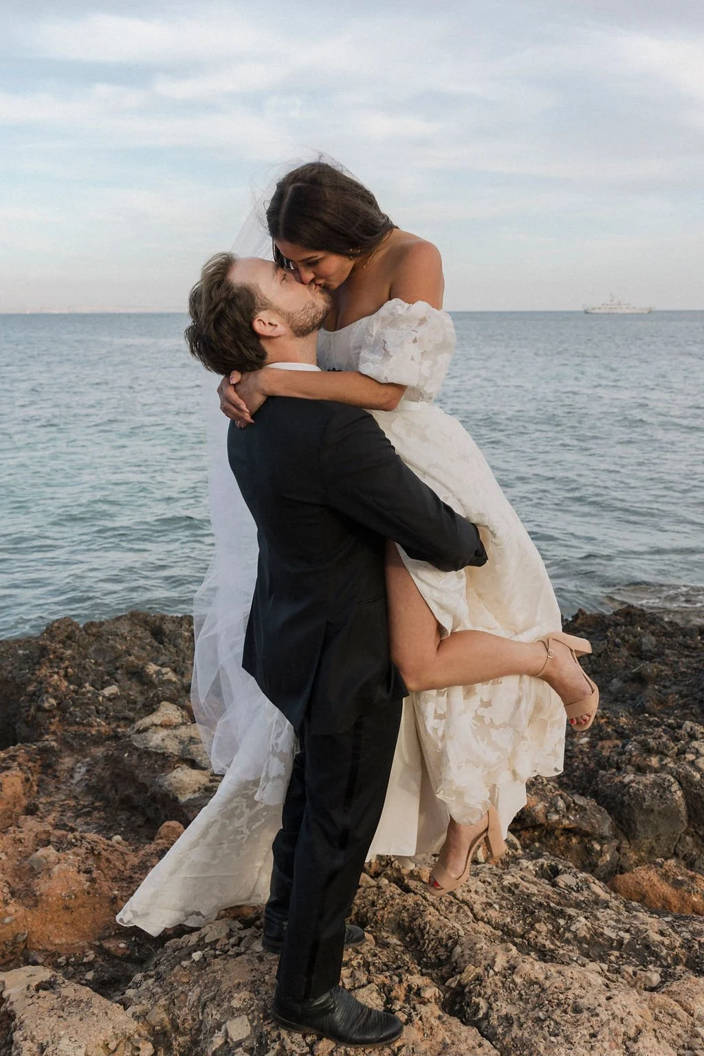 A groom lifting a bride in a wedding dress on rocky shoreline by the ocean, sharing a kiss with a boat in the background.