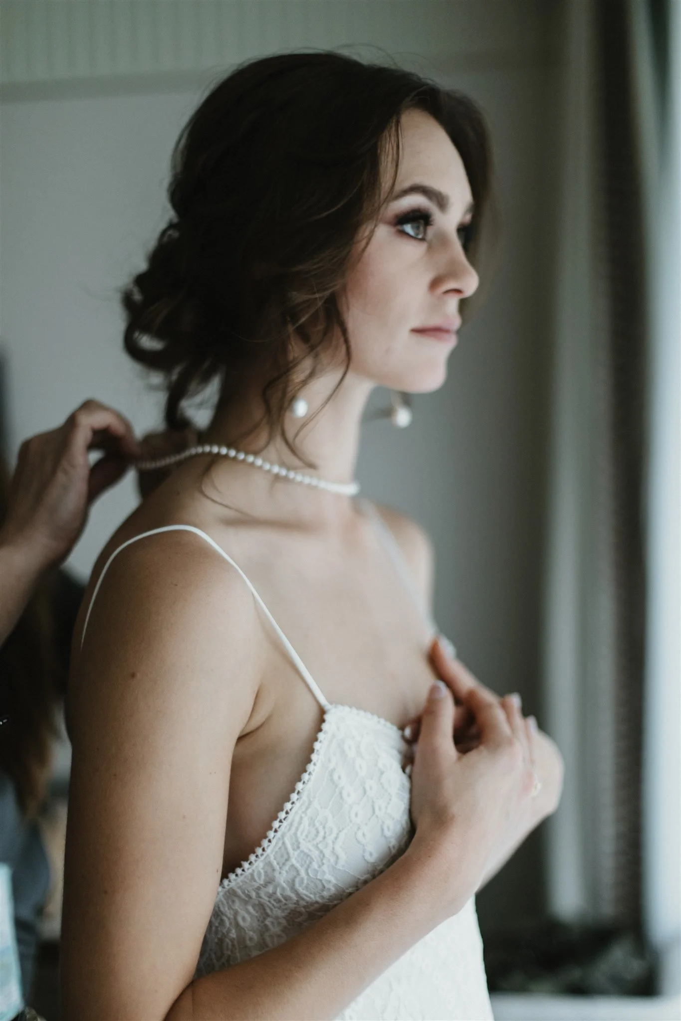A woman with brown hair, light skin, and blue eyes, wearing a white lace dress and a pearl necklace, is being helped with jewelry by a person whose hand is visible. The woman is looking to her right with a calm expression.