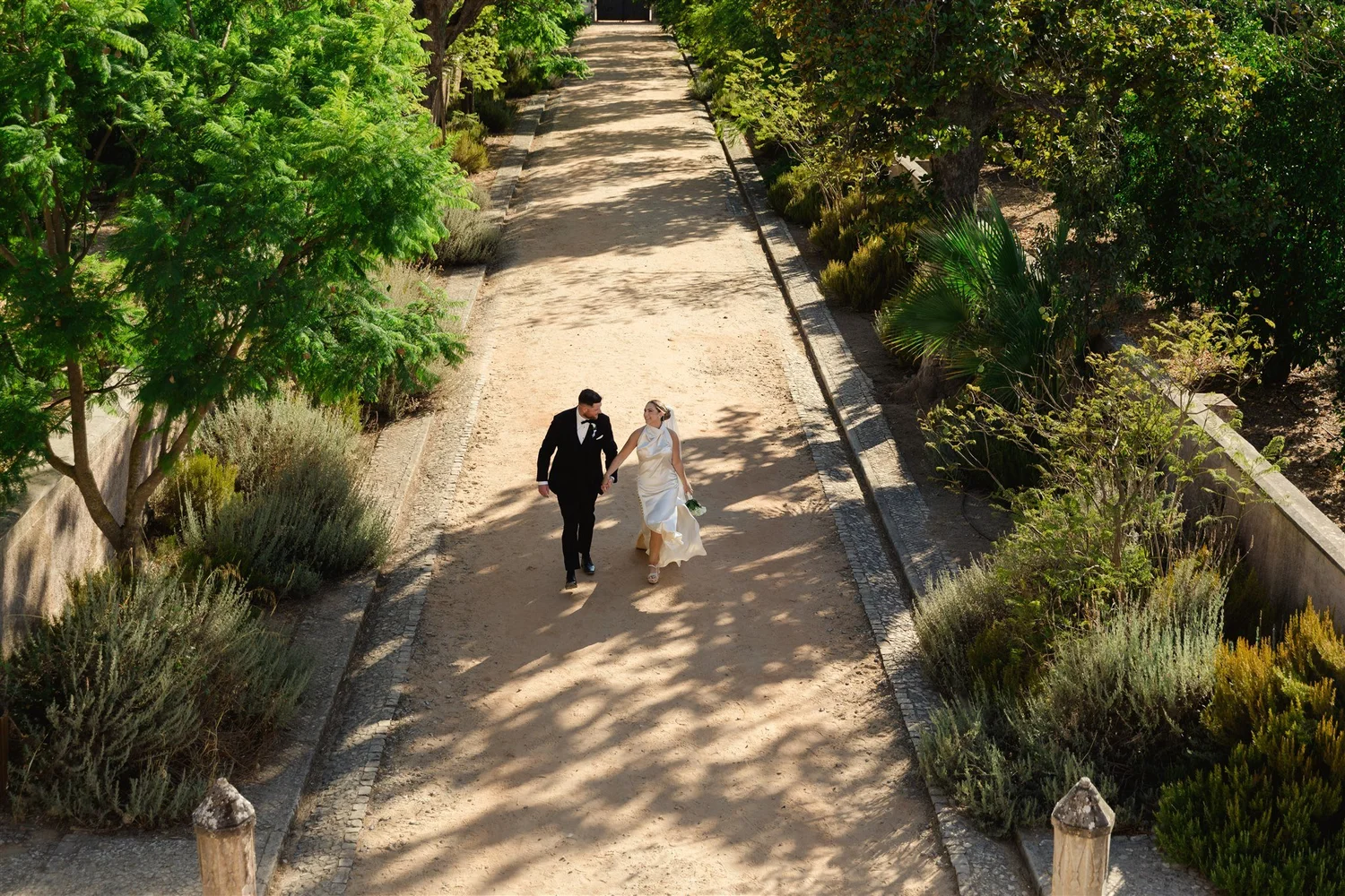A bride and groom walking hand in hand down a dirt pathway, surrounded by lush green trees and shrubs, casting dappled shadows on the ground.