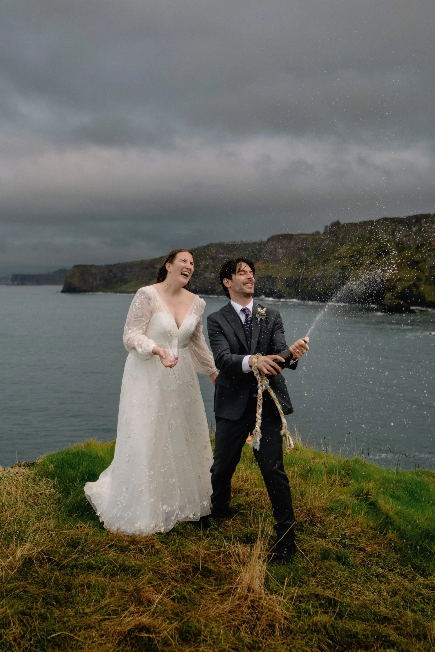 A bride and groom celebrating outdoors near water under a cloudy sky, with the groom spraying champagne and both laughing.