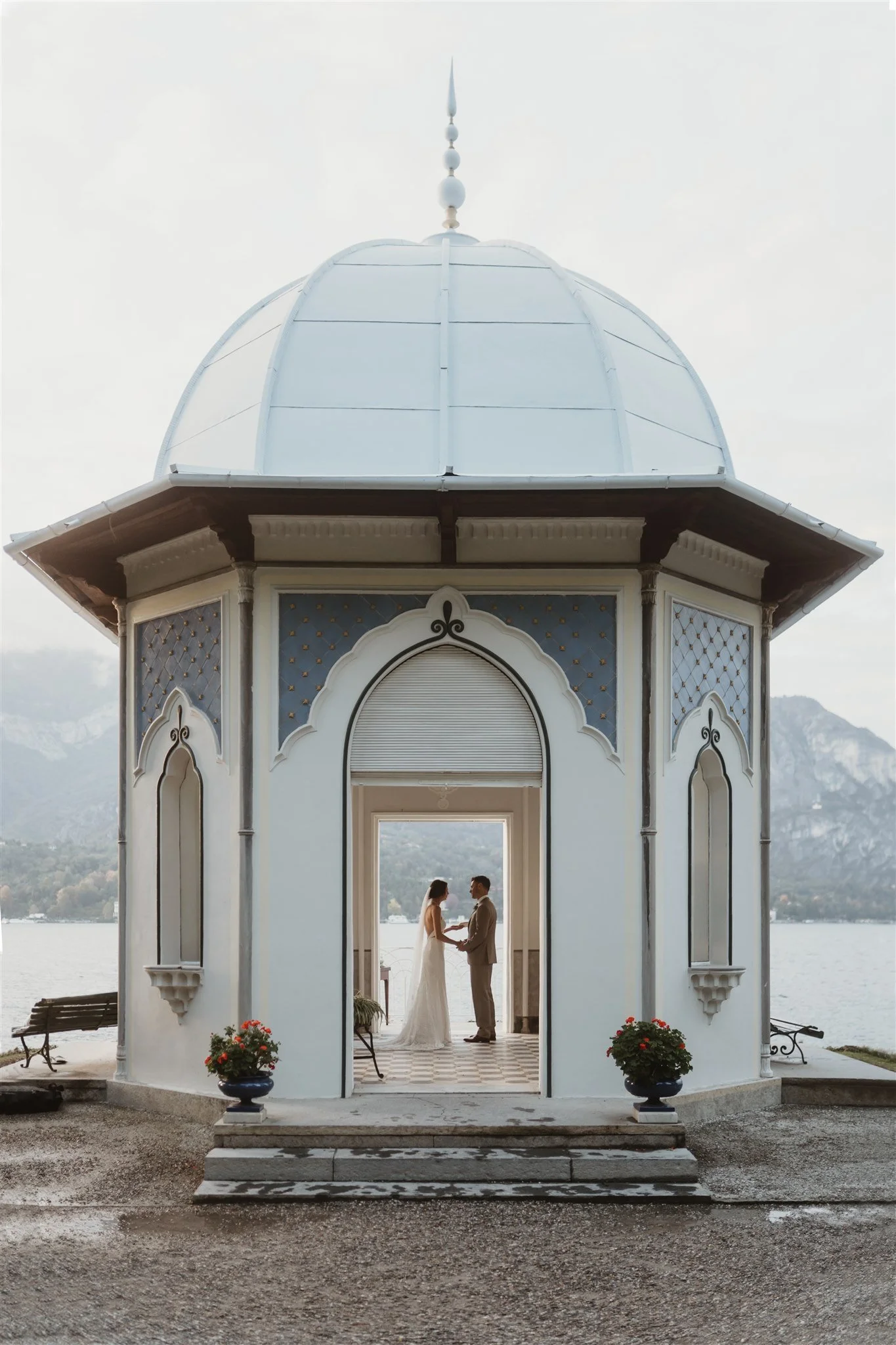 A couple stands holding hands inside a white decorative gazebo by the water, possibly during a wedding or engagement ceremony, with mountains in the background and potted plants at the entrance.