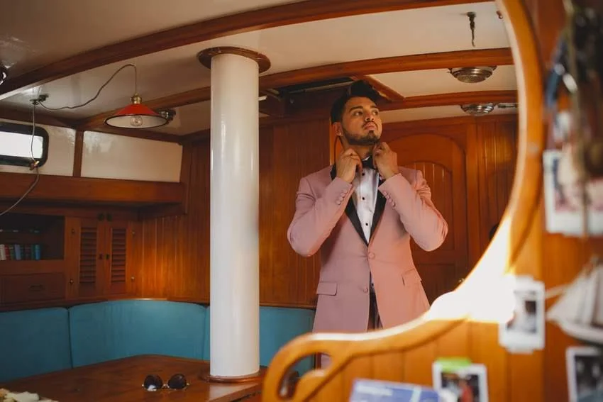 A man in a pink suit is adjusting his collar in front of a mirror, standing inside a wooden-paneled room.
