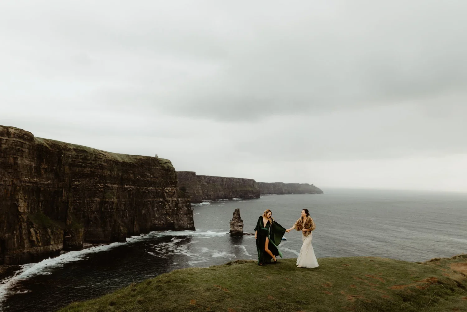 Two women in wedding attire standing near cliffs by the ocean, holding hands, with the cloudy sky overhead.