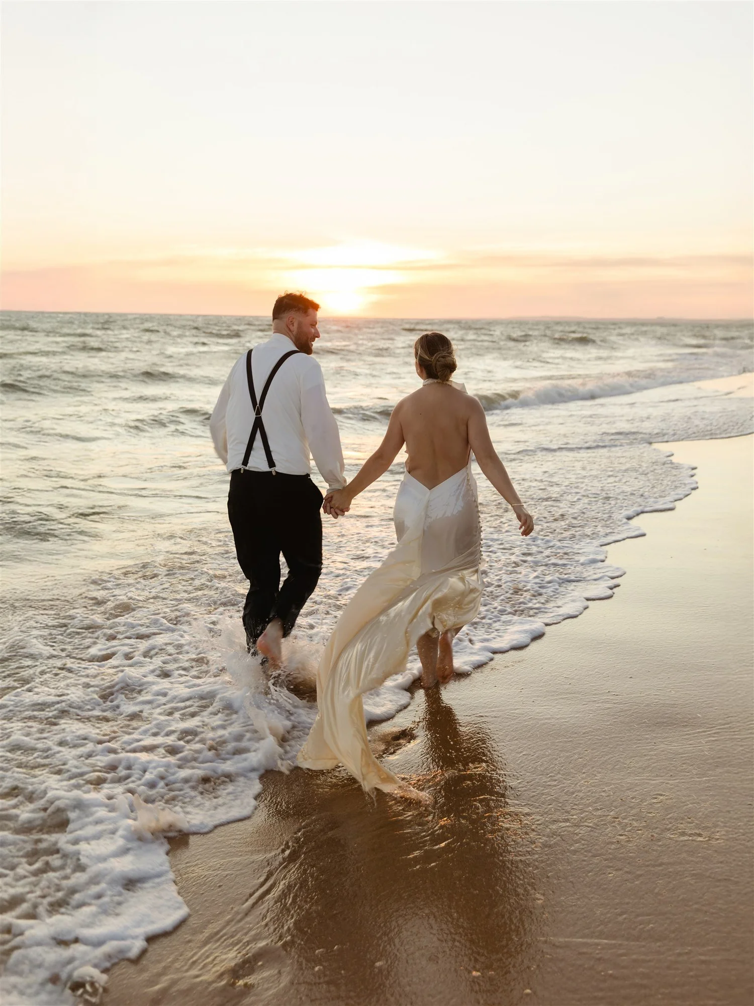 A couple dressed in wedding attire walking hand in hand along the shoreline at sunset, with waves lapping at their feet.