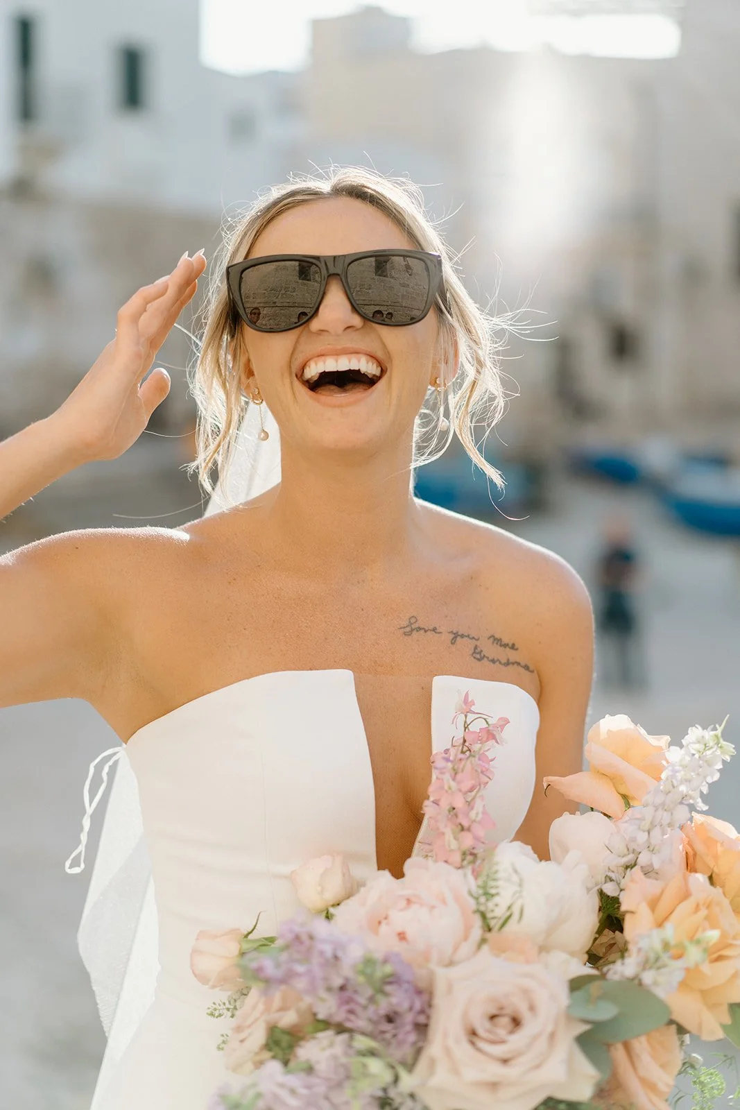 A smiling woman wearing sunglasses and a white strapless dress holding a bouquet of pink and white flowers outdoors, with blurred buildings in the background.