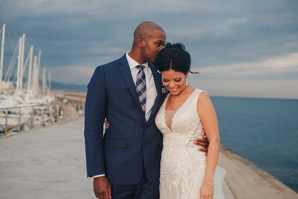 A newlywed couple stands close together on a dock by the water, with sailboats in the background. The groom, wearing a navy blue suit and plaid tie, kisses the bride on her forehead. The bride, in a lace wedding dress, smiles while looking down.