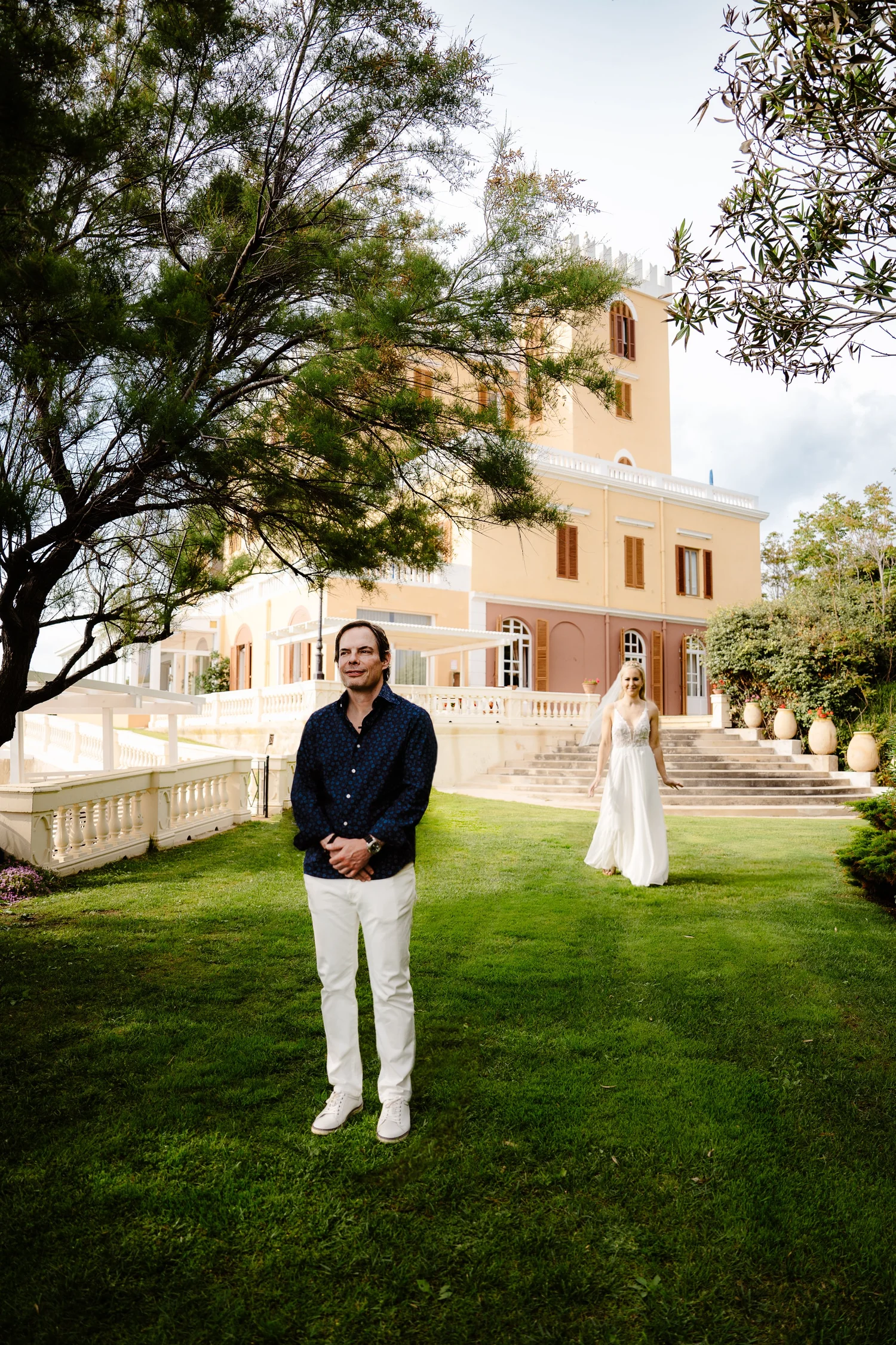 A man in white pants and a dark blue shirt stands on a lush green lawn, with a woman in a white dress and veil walking down stairs behind him. There is a large, peach-colored building with multiple stories and wooden shutters in the background, frame
