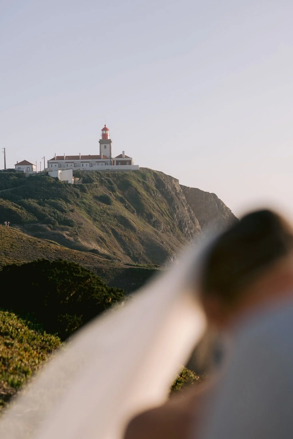 Elopement portrait of a lighthouse on a grassy cliff with a clear sky in the background, partially obscured by a blurred object in the foreground.