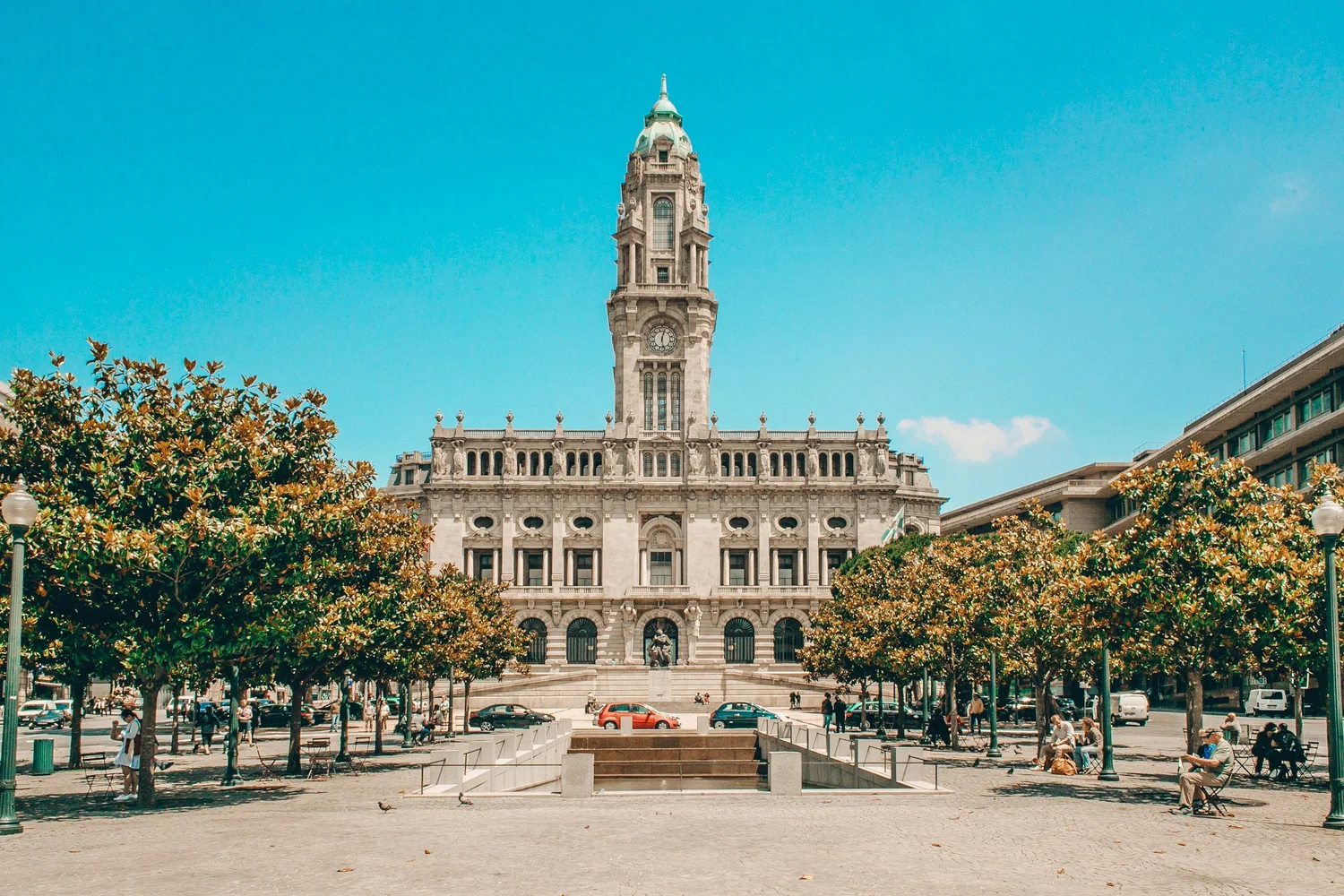 City square with people sitting on benches, trees, and cars parked along the street, in front of a large historic building with a clock tower.