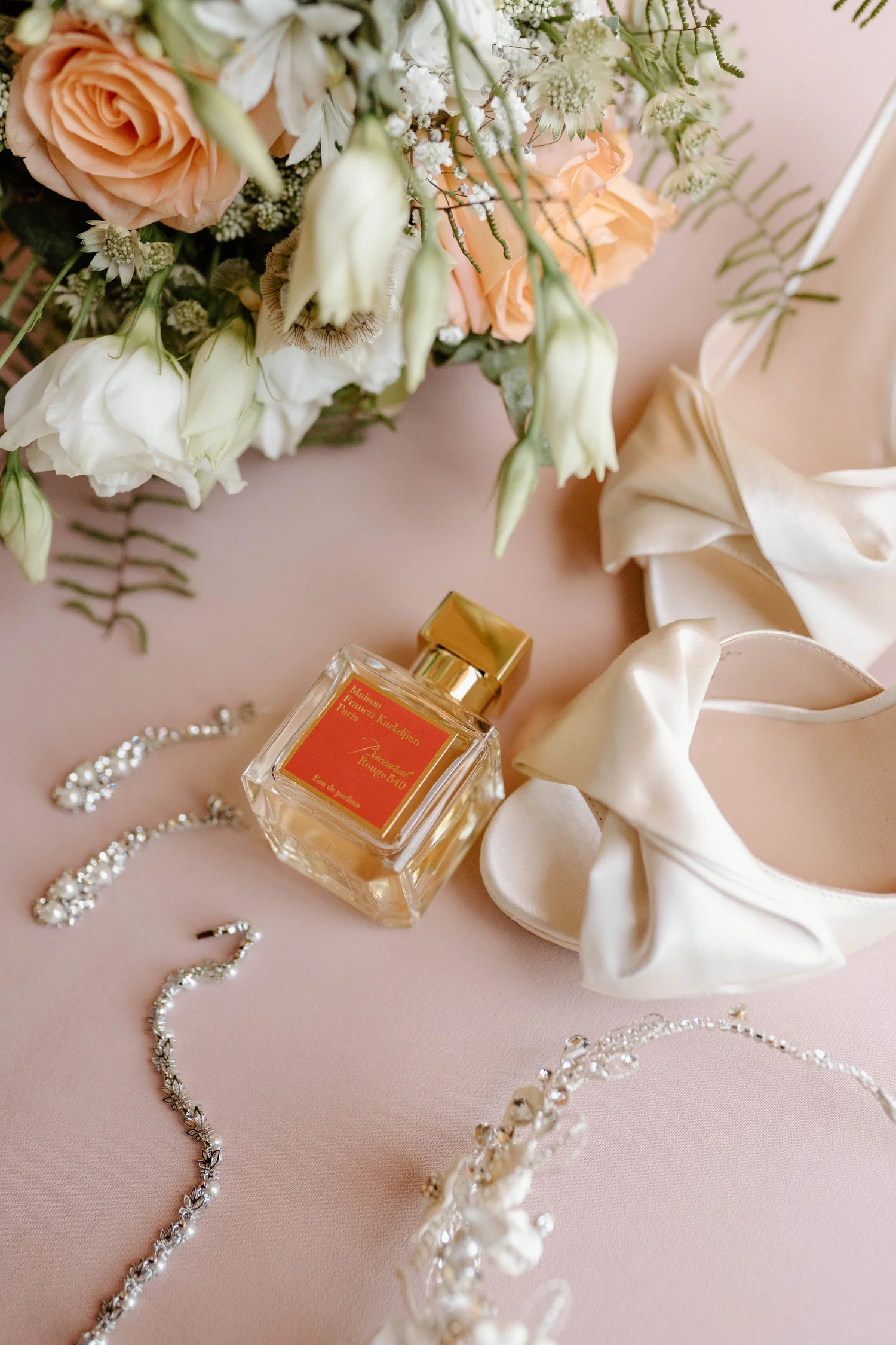 A floral bouquet with peach and white roses, white and green filler flowers, and greenery, placed on a pink background. Nearby are pearl and rhinestone necklaces, a cream-colored satin ribbon, a bottle of perfume, and a satin headband.