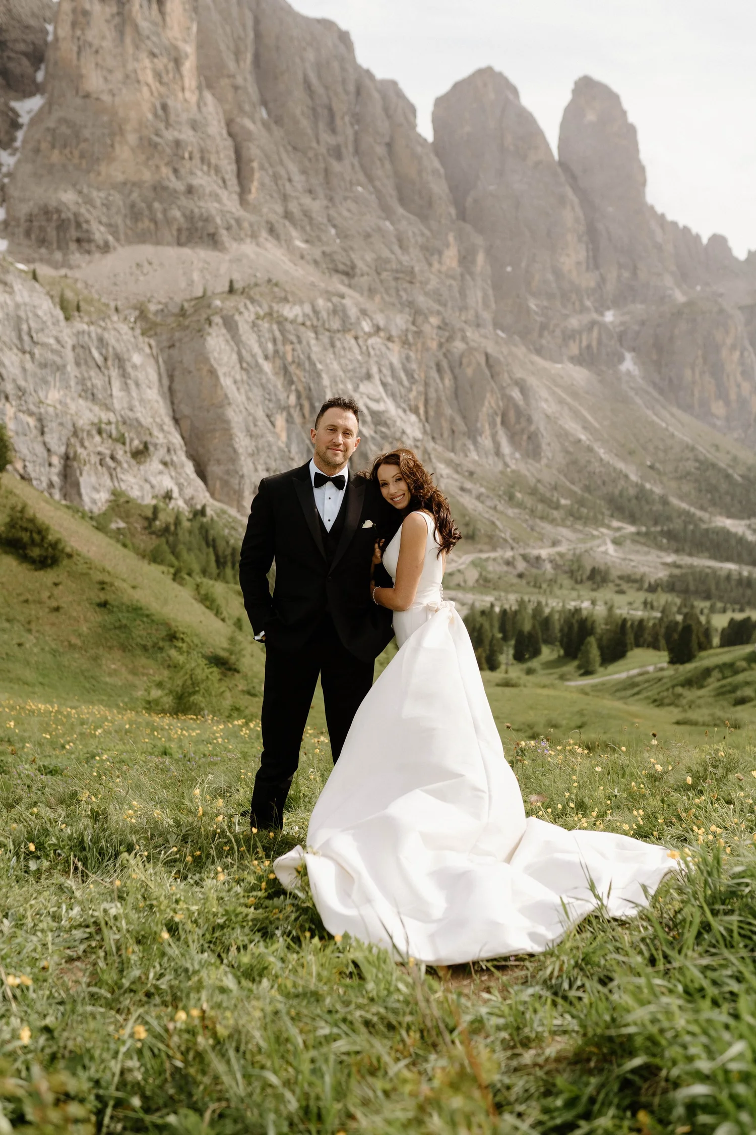 A newlywed couple in wedding attire standing on a grassy field with mountains in the background. The groom is in a black tuxedo and the bride is in a white gown with a train. They are smiling and embracing each other.