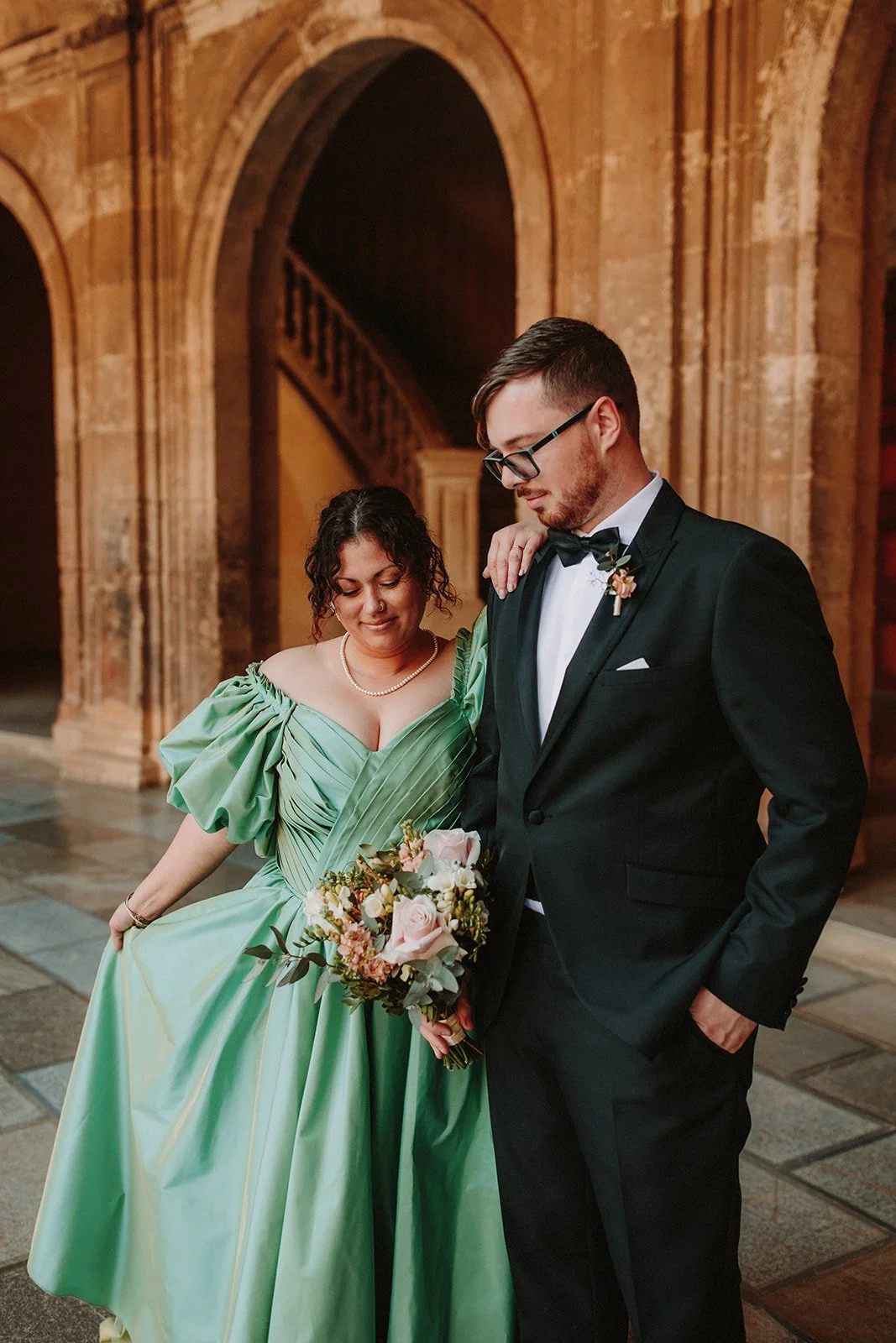 A couple dressed in formal wedding attire standing close together in an elegant, historic stone building. The woman is wearing a green gown with puffed sleeves and holding a bouquet, while the man is in a black tuxedo with a bow tie. They are looking down, with a romantic and tender moment.