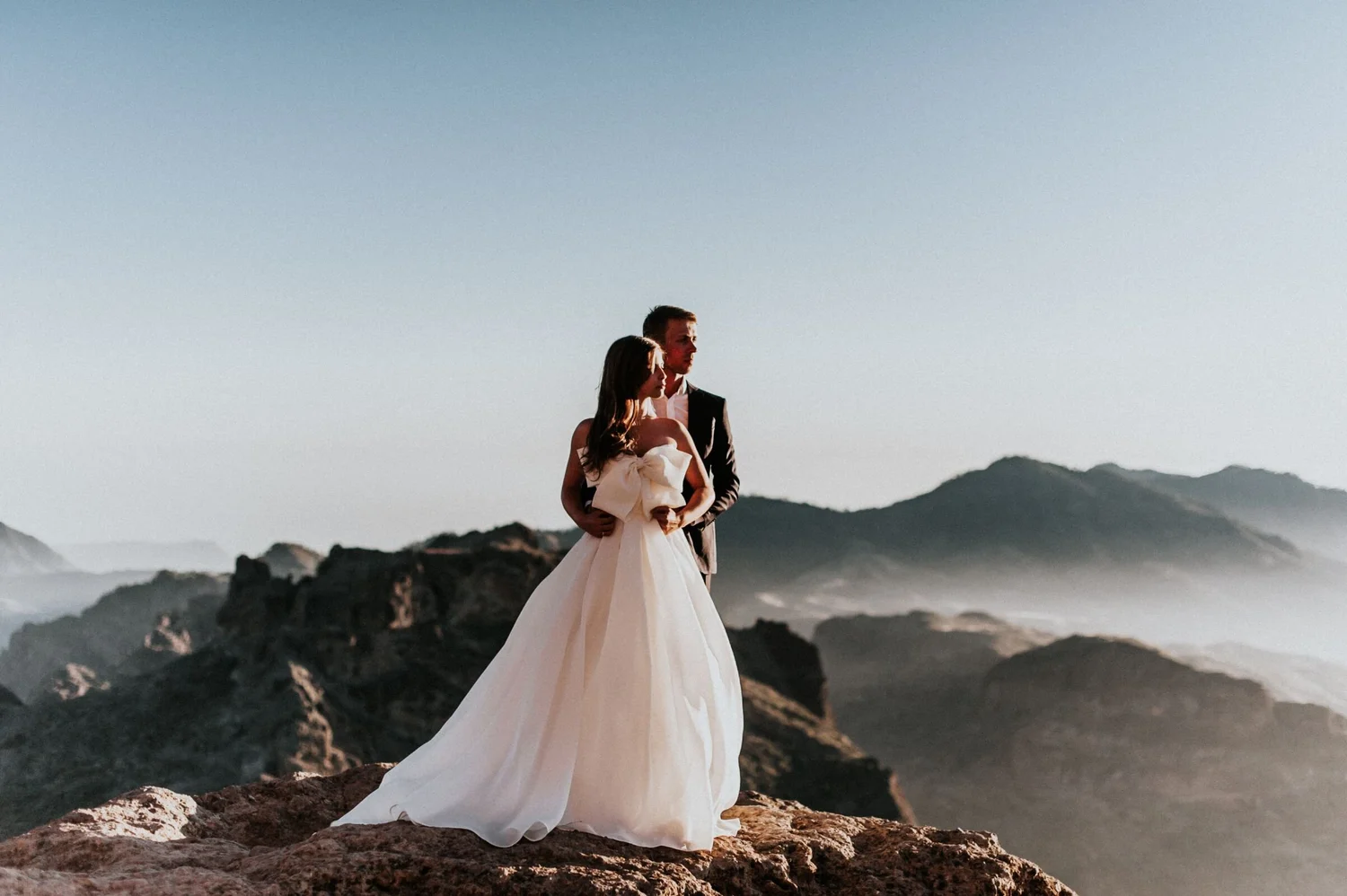 A bride and groom standing on rocky terrain outdoors during sunset, with mountains and sky in the background.