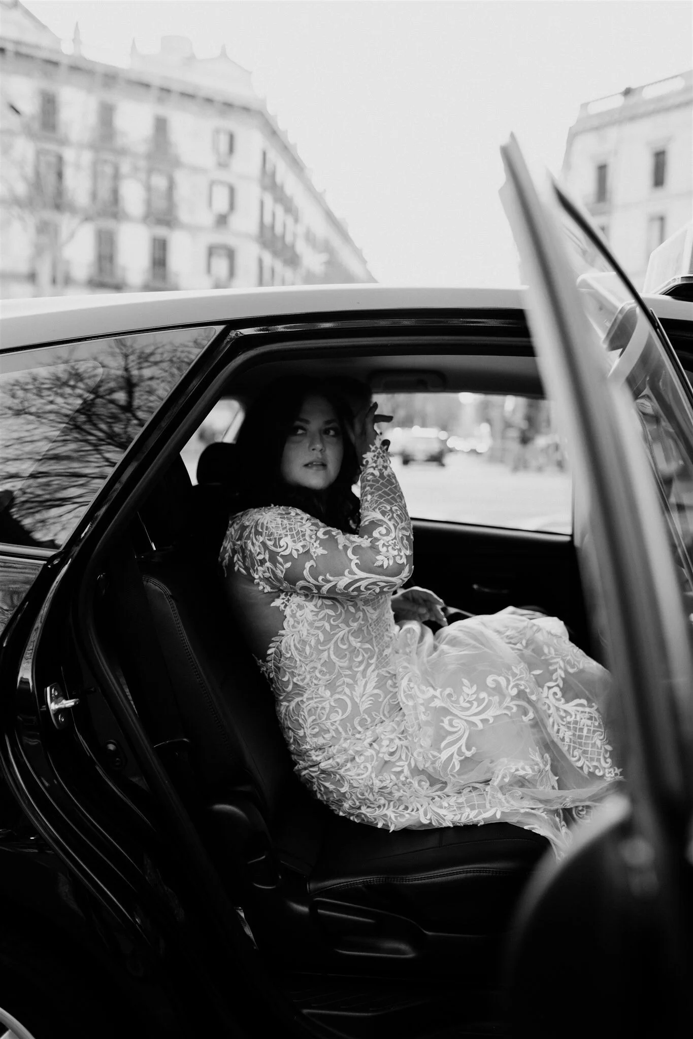 A woman in a lace dress sitting in the backseat of a car, raising her right arm and looking out the window. Buildings and a tree are visible outside.