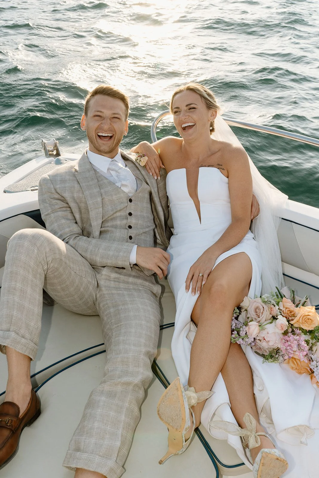 A happily married couple in wedding attire sitting on a boat, smiling and laughing with the ocean in the background, and a bouquet of flowers on the woman's lap.