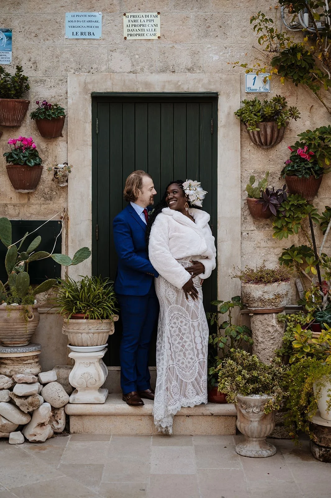 A couple dressed in wedding attire smiling and looking at each other, standing on a step surrounded by potted plants and flowers, in front of a green door with a stone frame.