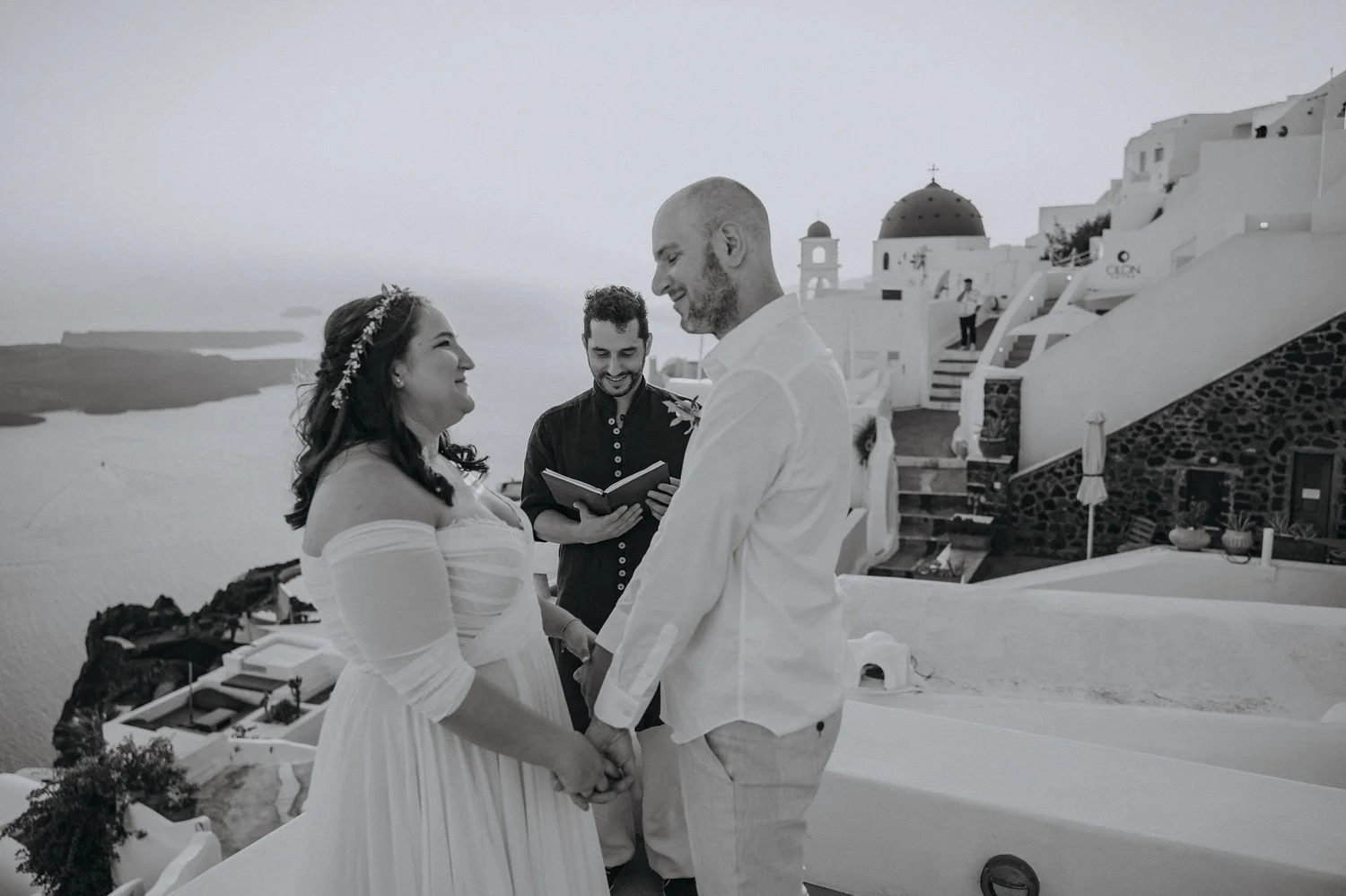 A couple holds hands and looks into each other's eyes during a wedding ceremony outdoors, with an officiant reading from a book in the background, in front of white buildings and stairs.