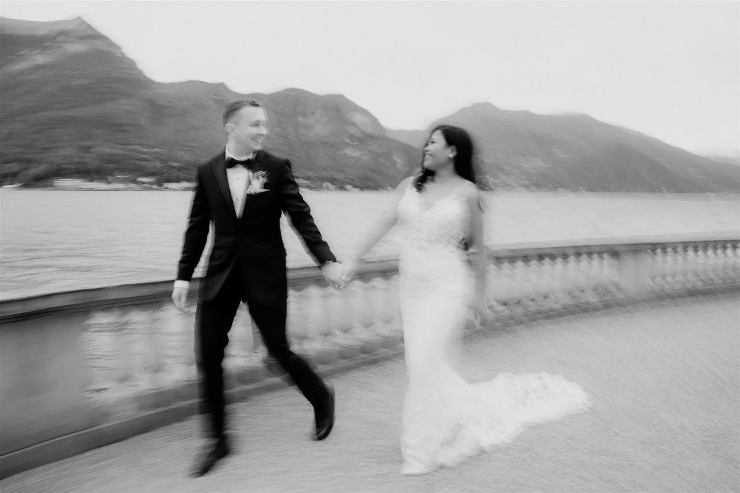 A couple holding hands, dressed in formal wedding attire, walking along a walkway near a body of water with mountains in the background.