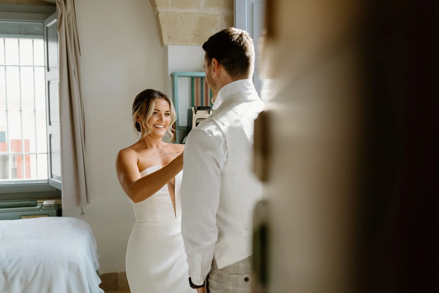 A woman in a white dress smiling at a man in a white shirt during a moment indoors, with a window and open window shutters in the background.