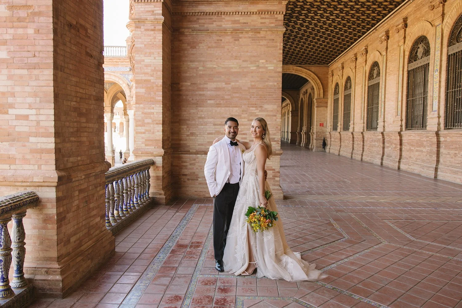 A newlywed couple standing on a brick balcony with ornate railings, dressed in wedding attire, with a historic building in the background.