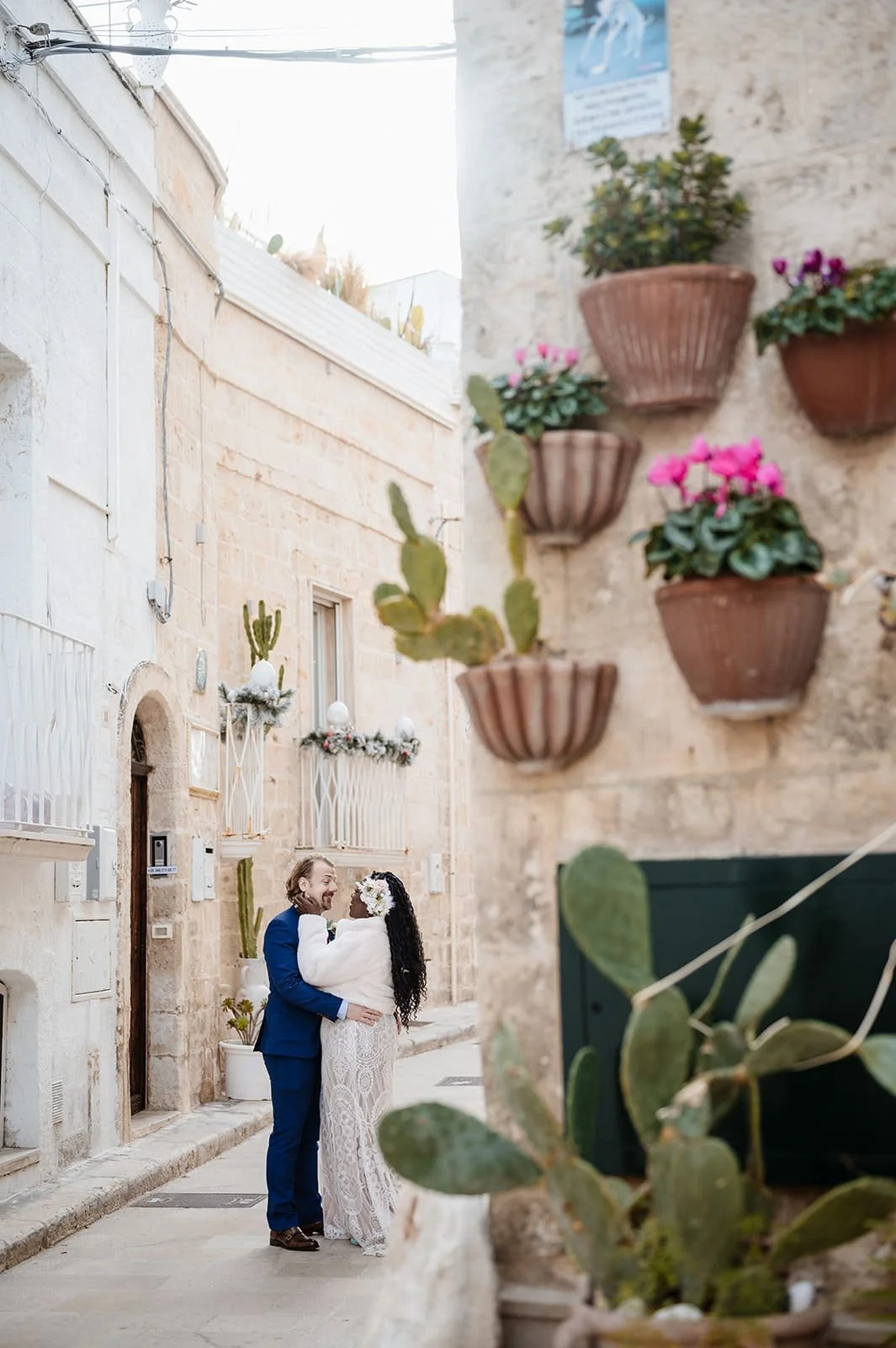 A couple dressed in formal attire, the man in a blue suit and the woman in a white lace dress, embracing and smiling at each other on a narrow street with beige stone buildings, potted plants, and decorative flowers.