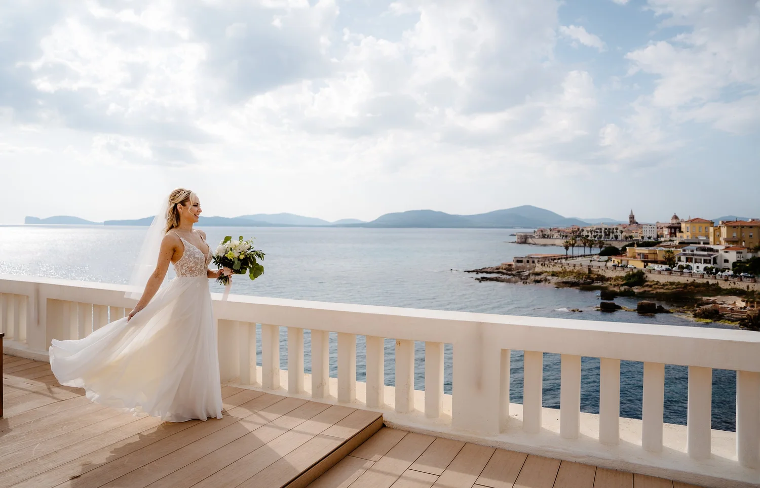 A smiling bride in a white wedding dress holding a bouquet of flowers, standing on a balcony overlooking the ocean and a seaside town with mountains in the background.