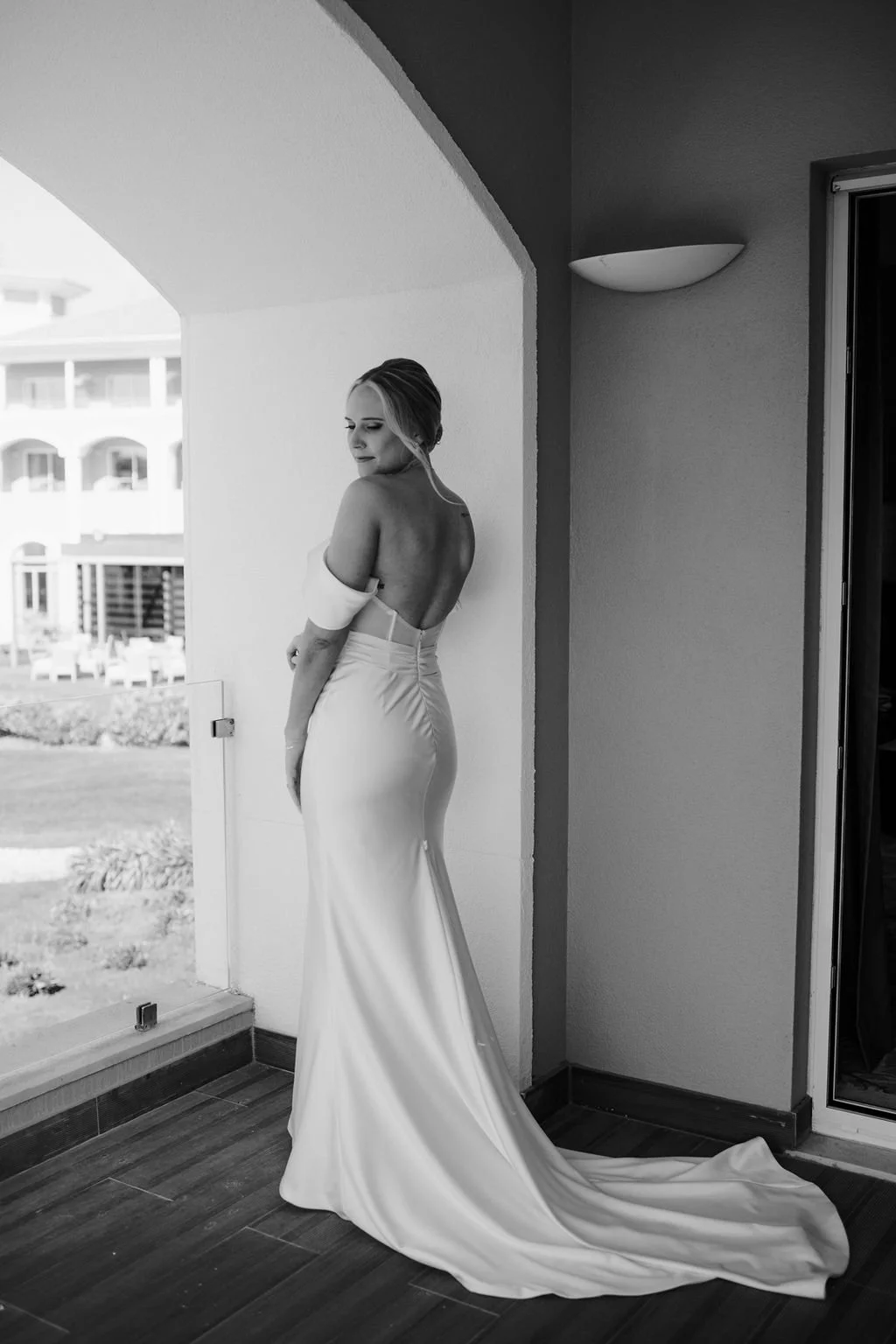 A woman in a backless, fitted wedding gown standing on a balcony, looking down with a serene expression.