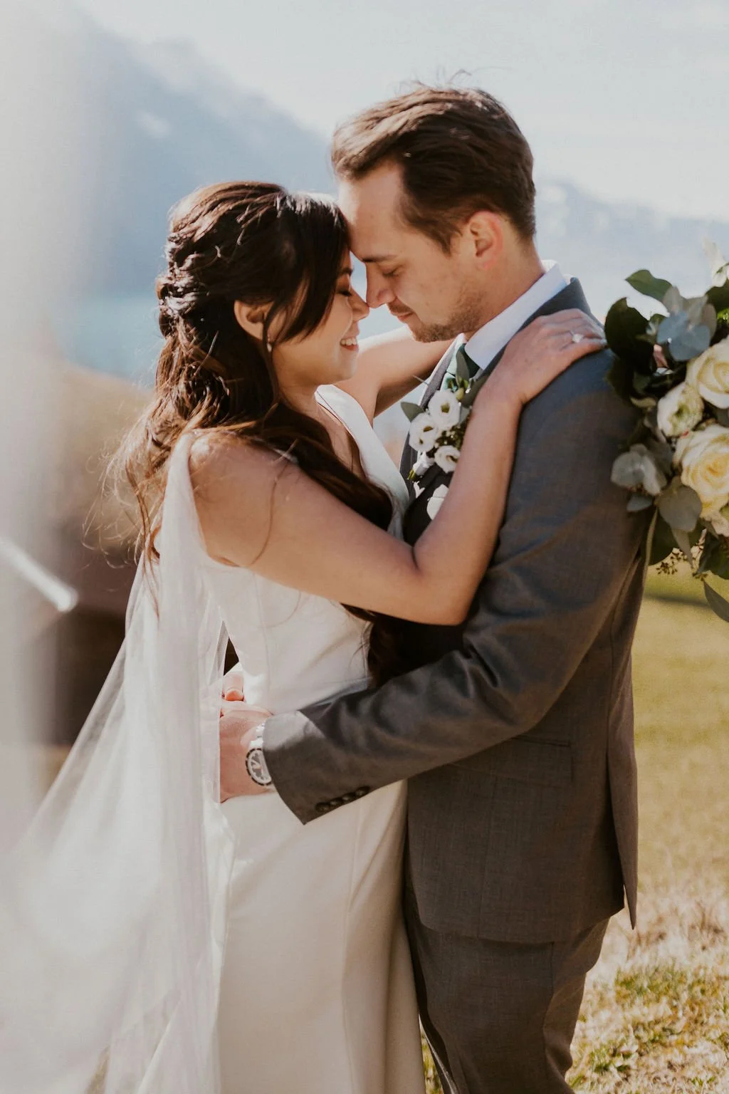 A bride and groom holding each other with foreheads touching outdoors during daytime, with mountains and a body of water in the background, celebrating their wedding.