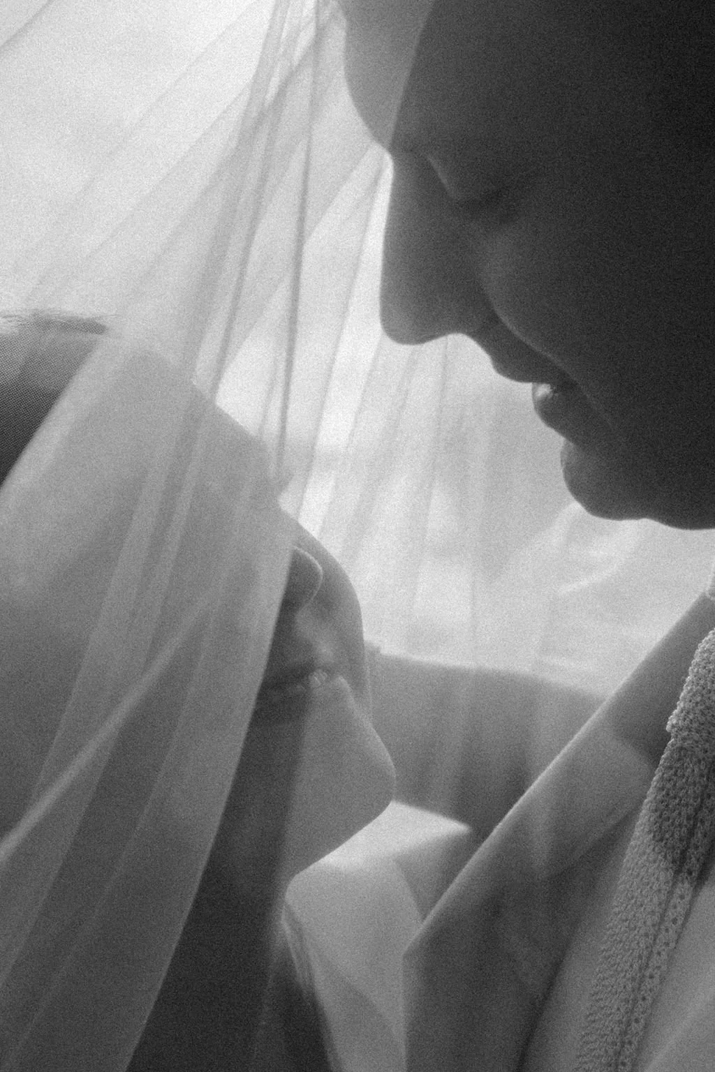 A black and white close-up photo of a young couple facing each other, separated by a semi-transparent curtain, with their faces closely approaching each other.