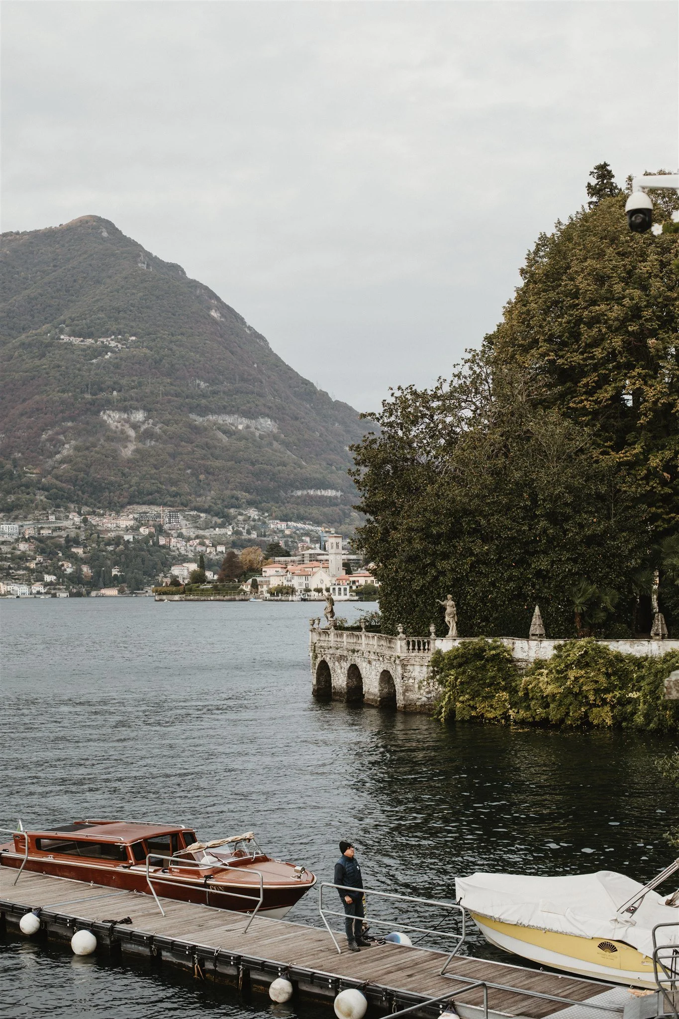 A lakeside scene with boats docked at a pier, a man walking on the dock, trees, a castle-like structure on a hill, and a mountain in the background.