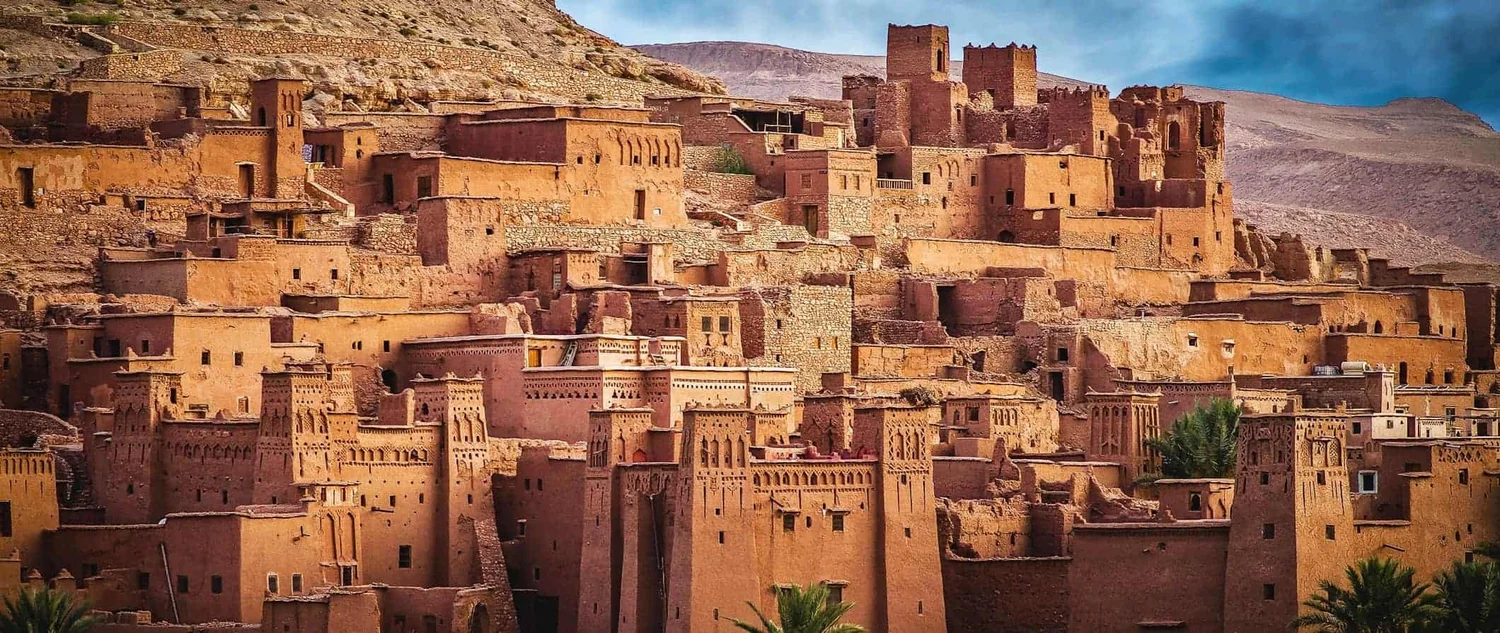 Traditional desert village made of mud bricks, built on a hillside, featuring multiple layered structures and towers, with mountains in the background.