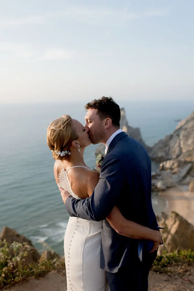 A bride and groom sharing a kiss on a beach at sunset, with rocky cliffs and the ocean in the background.