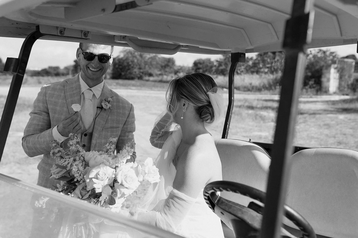A man in sunglasses and a checkered suit holding a flower bouquet stands next to a woman sitting in a golf cart, wearing a wedding dress and veil. The man is smiling and the woman is looking down, with a bouquet of flowers in front of her. They are o