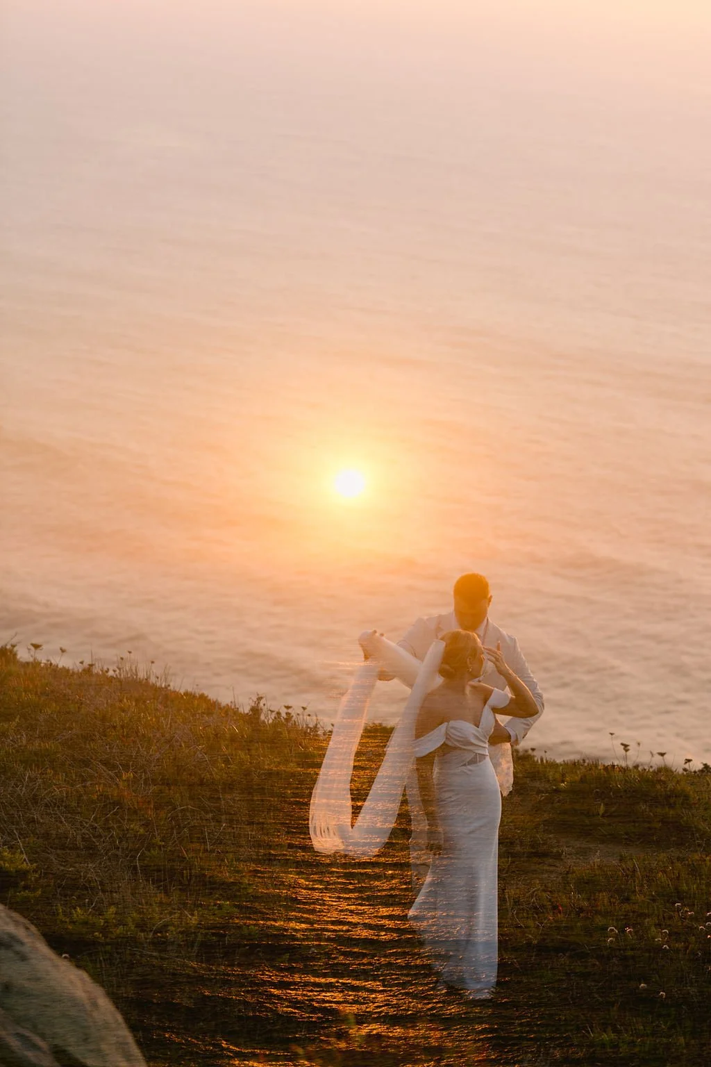 A couple embracing and kissing by the water at sunset, with a semi-transparent overlay of the same couple in a romantic pose.