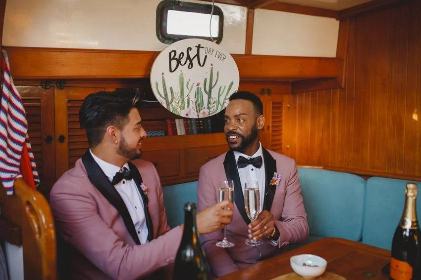 Two men in pink tuxedos and black bow ties sitting in a wooden booth with blue seating, holding champagne glasses, celebrating together. One man has a beard, the other is clean-shaven. There is a bottle and a small bowl on the table. Behind them, a r