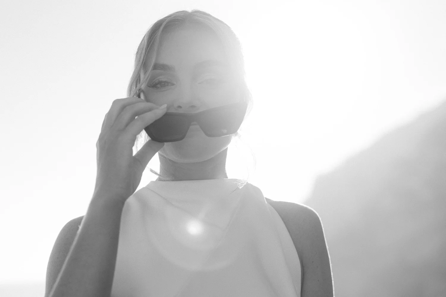 A woman is holding a camera close to her face, partially covering her mouth with her hand, and looking at the camera. She is outdoors with a bright, overexposed background.