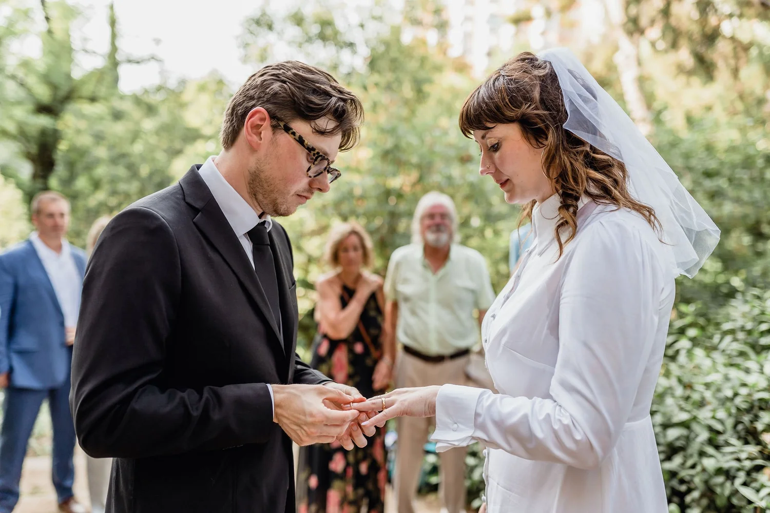 A couple exchanging wedding vows outdoors, with a small group of friends and family watching in the background.