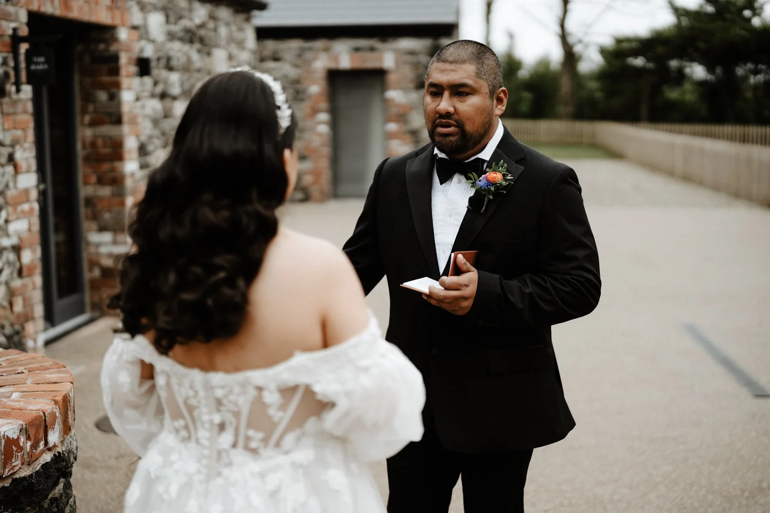 A man in a tuxedo with a boutonniere is holding a small box and a card, looking at a woman in a white lace dress with off-shoulder sleeves, standing outside near a brick wall.
