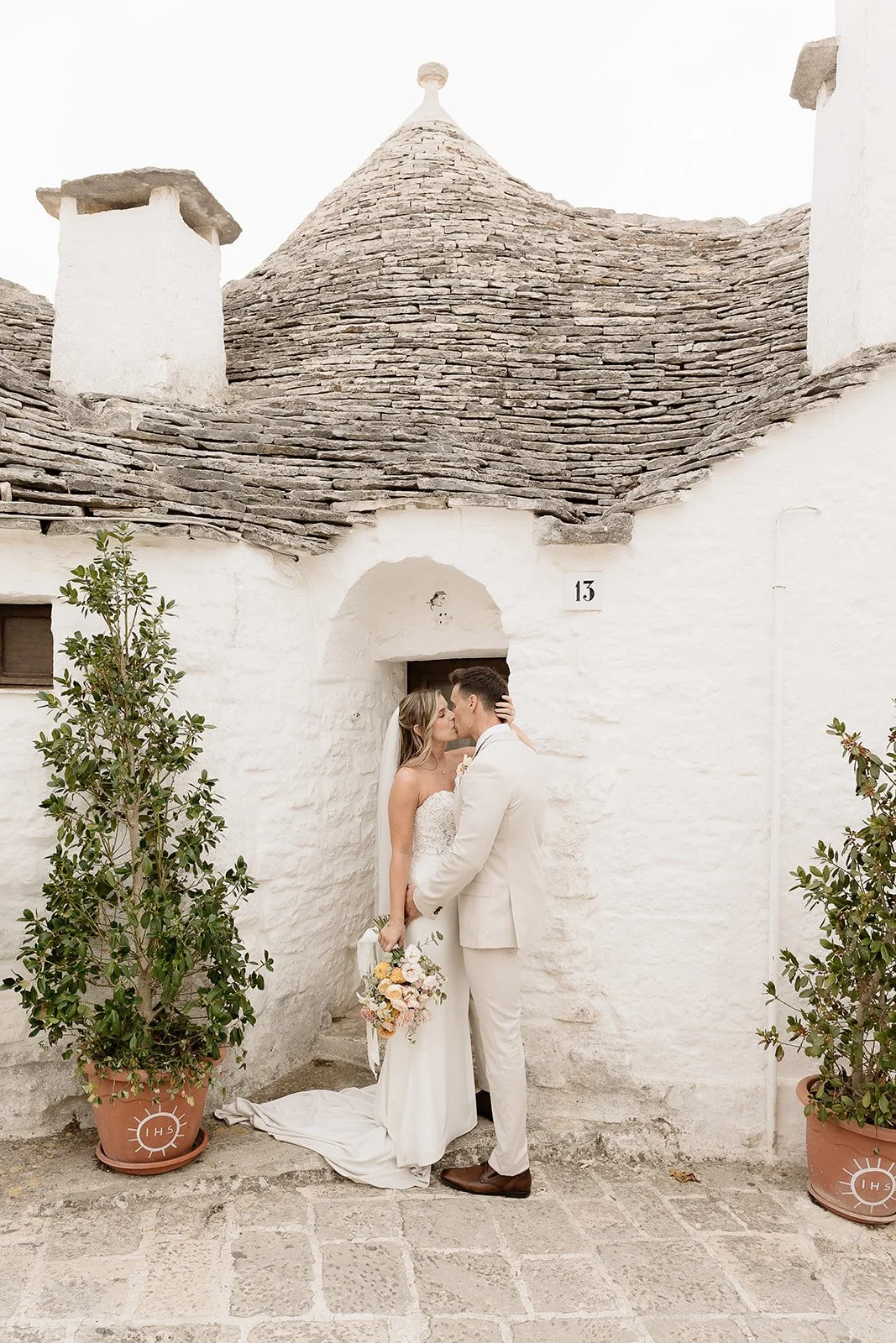 A bride and groom kiss in front of a white stone building with a conical roof, two potted plants, and the number 13 on the wall.