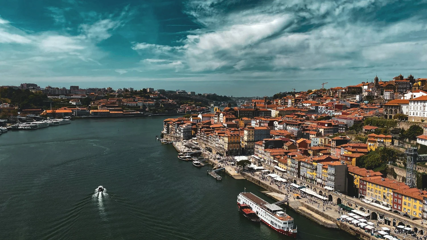 A picturesque view of a city with colorful buildings along a river, with boats docked and sailing, and a hilly backdrop under a partly cloudy sky.