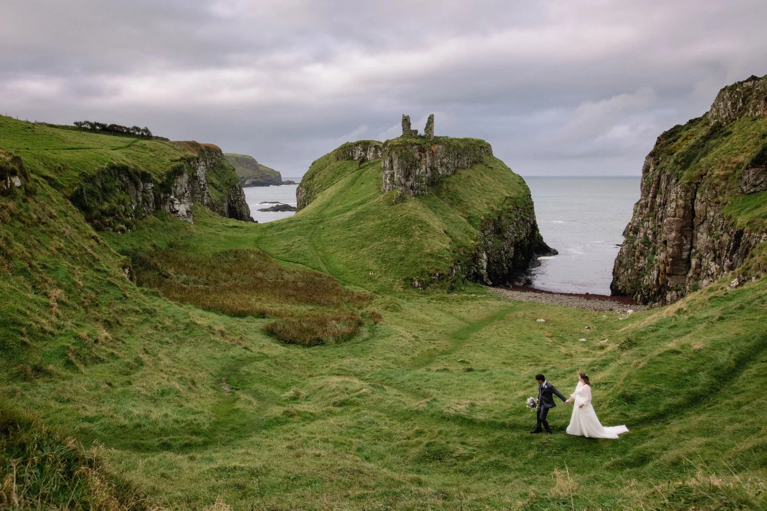 A bride and groom holding hands and walking across a grassy field near cliffs with ruins on top, overlooking the ocean under an overcast sky.