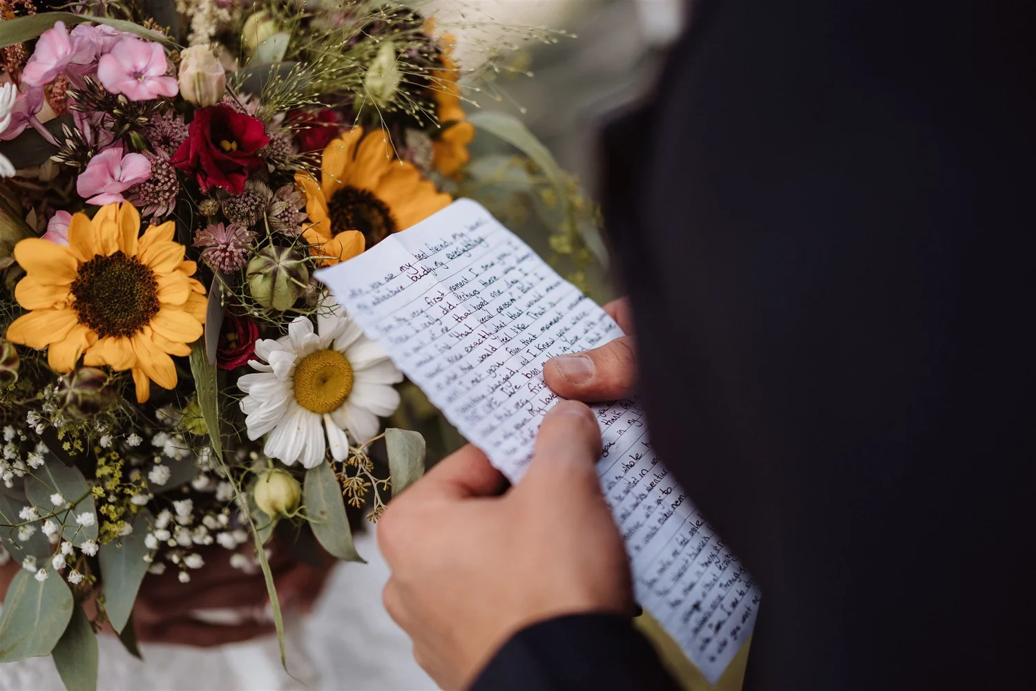 A person holding a handwritten letter near a bouquet of flowers, including sunflowers, daisies, and baby's breath.